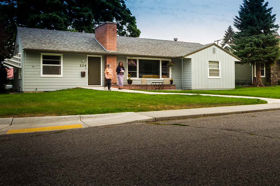 Two women, one with a flower-print skirt, the other a flower-print top, walk along a natural curving sidewalk away from a one-story bluish gray house with a red brick chimney. The path goes through a grassy lawn and connects to a passing sidewalk, which has a curb-ramp that leads to the street.